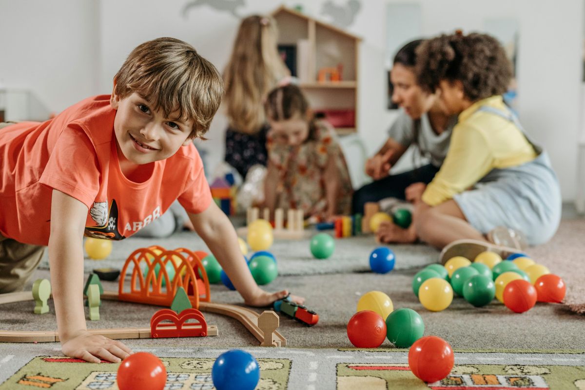 Ein Junge schaut in die Kamera, während er mit Spielzeugen spielt. Im Hintergrund sind weitere Kinder und eine Frau.
